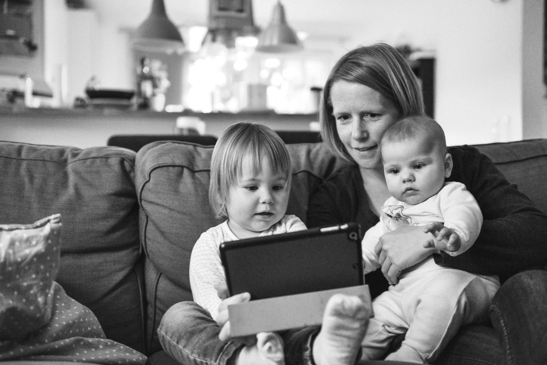 A parent and two young children looking at a tablet together on the sofa
